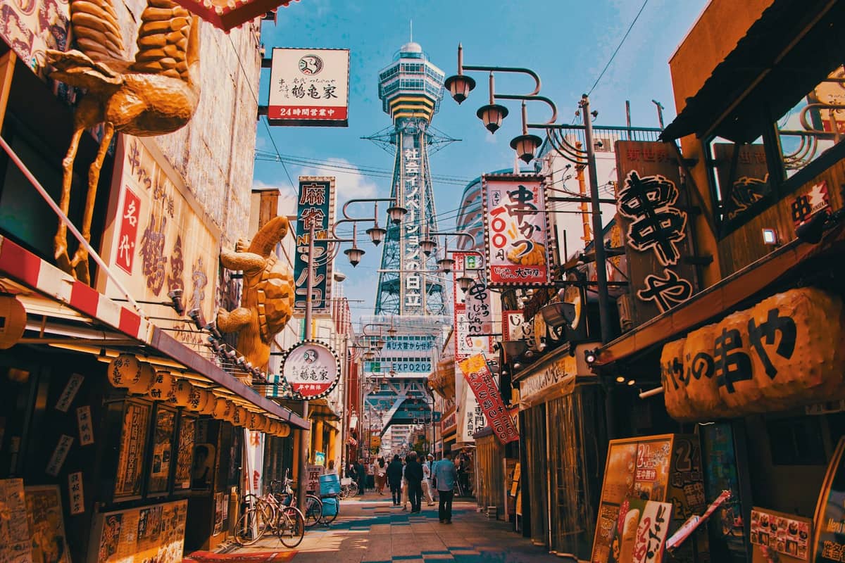 The colorful main street of Shinsekai in Osaka, with neon signs, kushikatsu restaurants, and Tsutenkaku tower rising in the background on a sunny day.