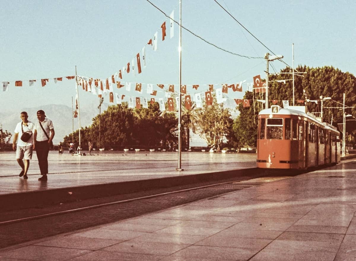 Antalya nostalgic tram at a city square with Turkish flags overhead and people walking nearby.