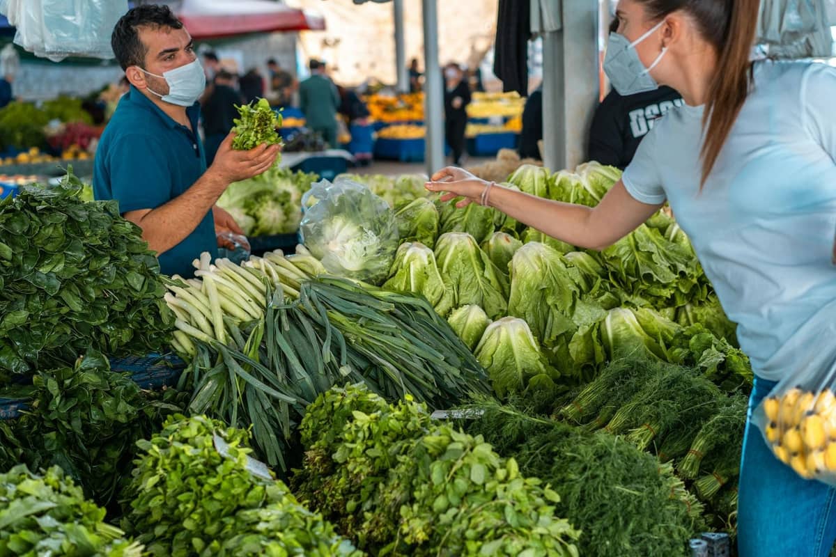 Shoppers wearing masks browse fresh greens and vegetables at a crowded outdoor market stall in Antalya.