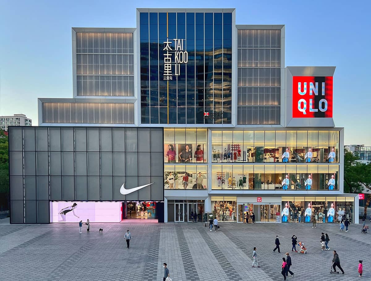 Moderne Fassade des Taikoo Li Einkaufszentrums in Peking in der Abenddämmerung mit Nike- und Uniqlo-Flagship-Stores, Glasfenstern und Passanten vor dem Gebäude.