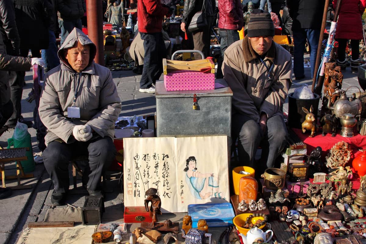 Zwei Verkäufer in Winterkleidung sitzen hinter einer Auslage mit Antiquitäten, Kunst und Nippsachen auf einem Außenmarkt in Beijing, mit Käufern im Hintergrund.