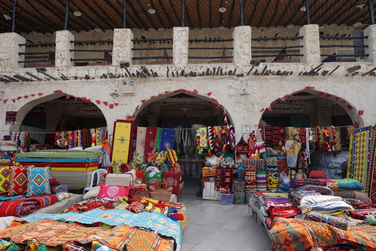 Teppich- und Stoffmarkt im Souq Waqif in Doha mit Bogensteinwerk und bunten Textilien vor traditionellen Läden.