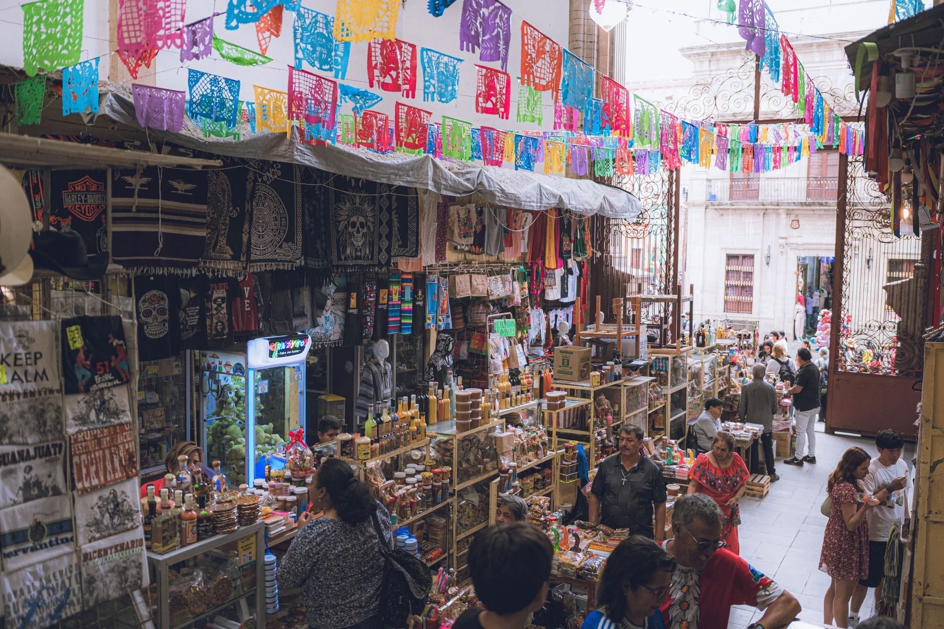 Mercado coberto em Guadalajara cheio de artesanatos coloridos, papel picado decorando o teto e pessoas fazendo compras nas barracas dos vendedores.
