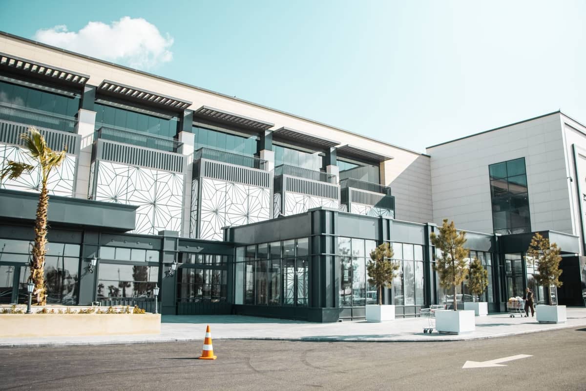 Modernes Einkaufszentrum-Außengebäude mit großen Fenstern, geometrischen Mustern, Palmen und einem sauberen, weitläufigen Eingang unter blauem Himmel.