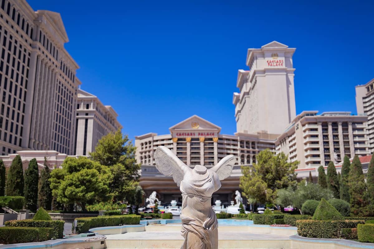 วิวกว้างของด้านนอกรีสอร์ท Caesars Palace ในลาสเวกัส พร้อมรูปปั้นสไตล์โรมัน ต้นไม้สีเขียว และท้องฟ้าสีฟ้าใส