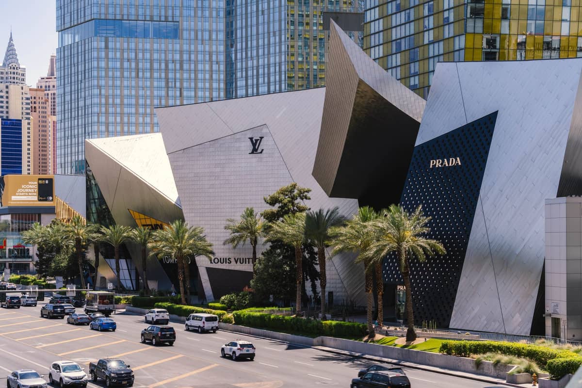 Wide landscape view of Las Vegas Strip showing upscale shopping at The Shops at Crystals with Prada and Louis Vuitton storefronts and palm trees lining the street.