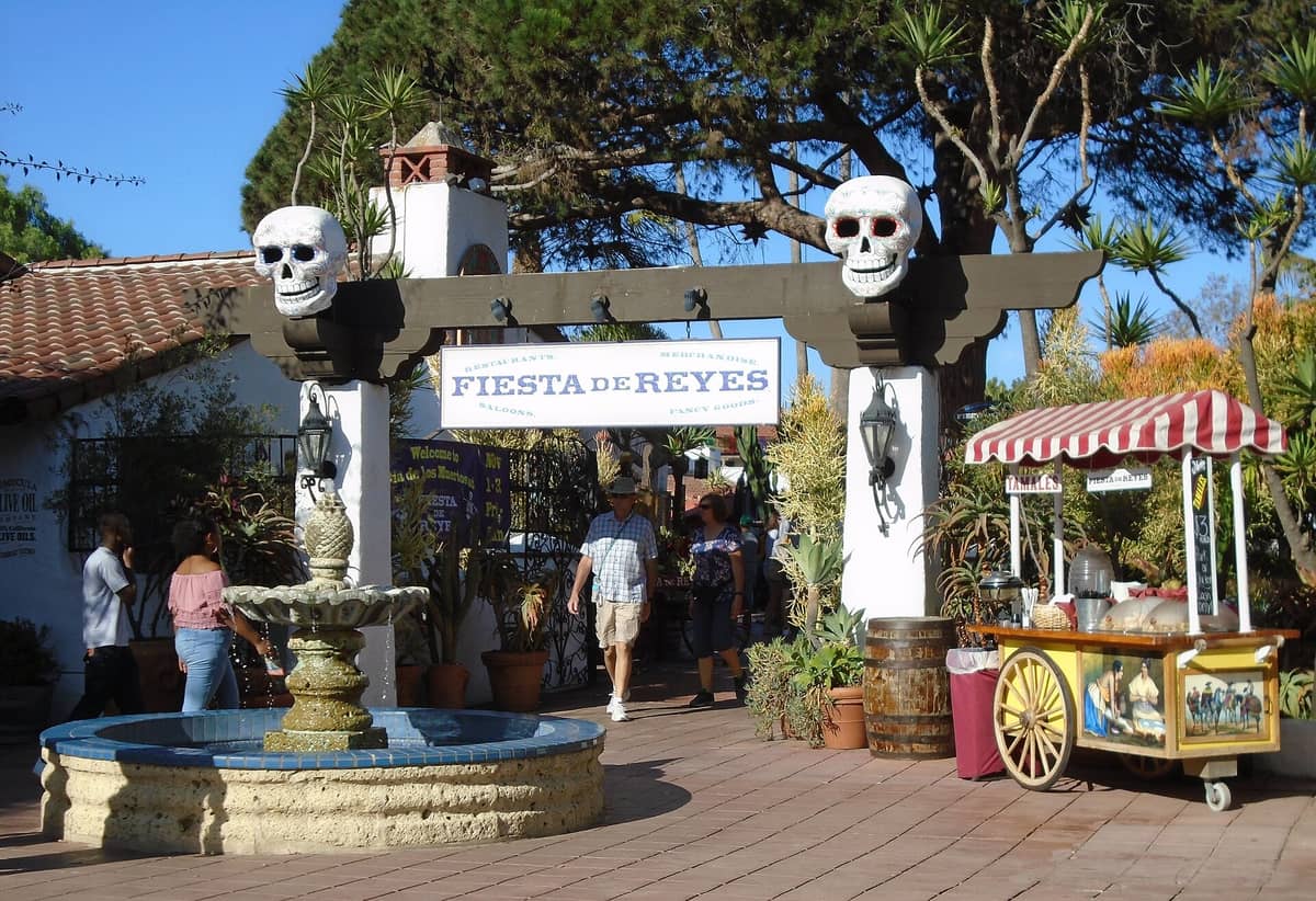 Eingang zum Fiesta de Reyes im Old Town San Diego mit einem dekorativen Schild, Totenköpfen, Brunnen, Essensstand und einkaufenden Menschen.