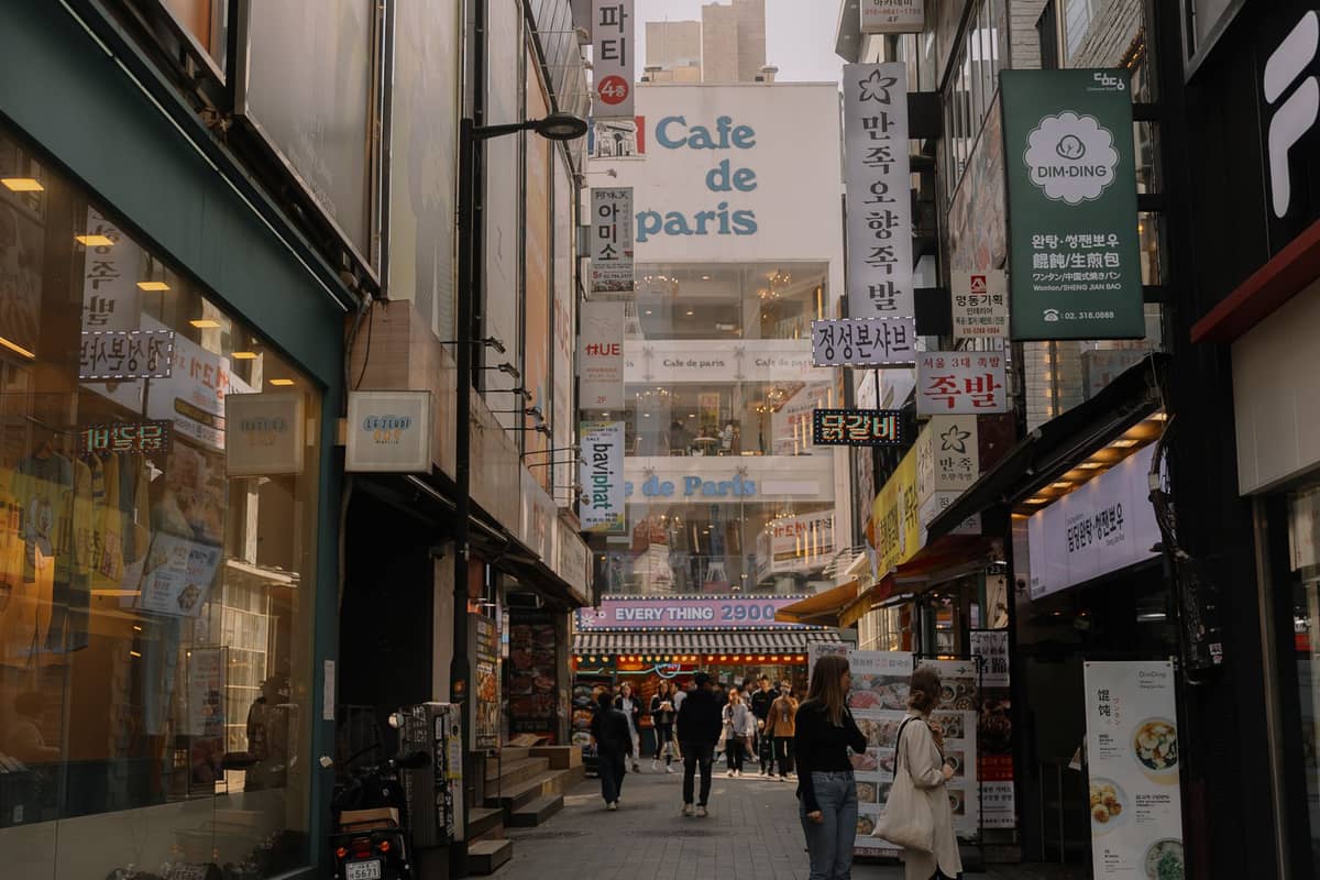 Uma movimentada viela urbana em Seul repleta de lojas, restaurantes e letreiros coreanos coloridos, cheia de pessoas e com uma atmosfera urbana vibrante.