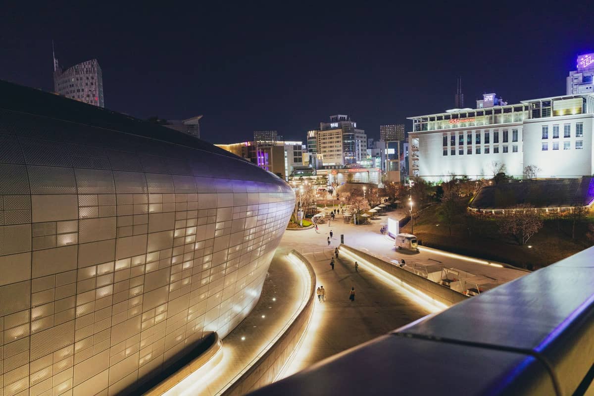 Vista noturna da Dongdaemun Design Plaza com passarelas iluminadas, pessoas e edifícios comerciais ao redor em Seul.