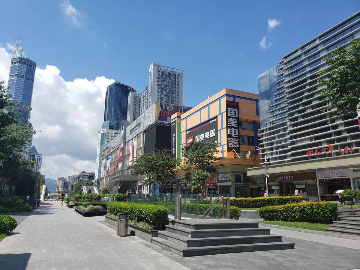 Ampla avenida de pedestres em Shenzhen ladeada por shoppings modernos, lojas e arranha-céus sob um céu azul e ensolarado.