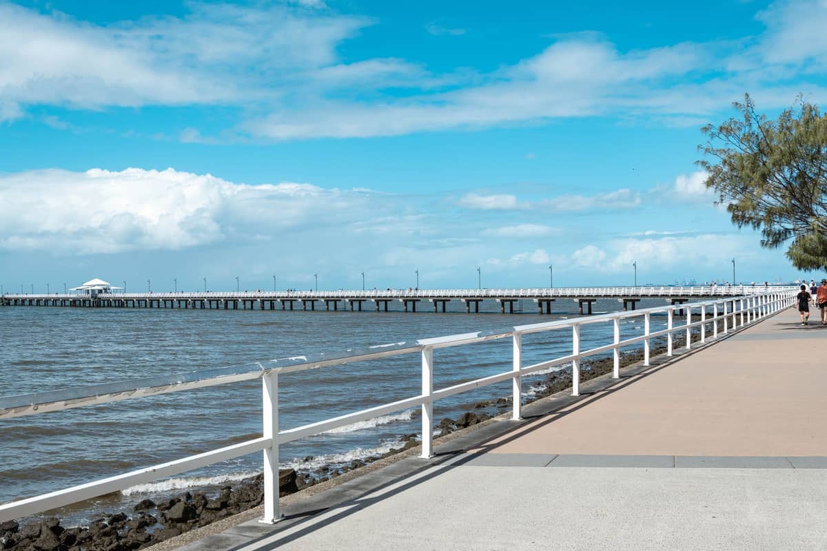 Der lange Holzsteg des Shorncliffe Pier erstreckt sich unter strahlend blauem Himmel über die Moreton Bay, während Menschen auf der Uferpromenade flanieren.