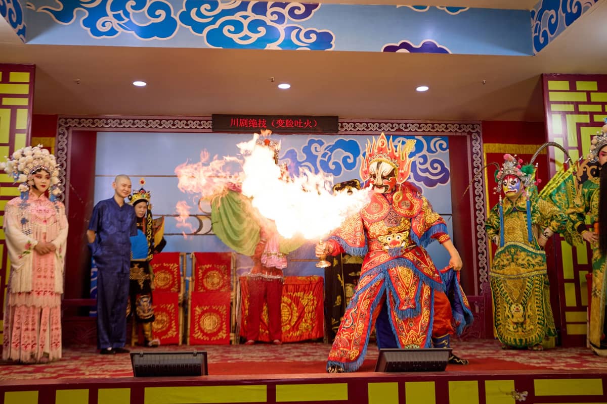 Darsteller der Sichuan-Oper auf der Bühne in bunten traditionellen Kostümen; ein zentraler Darsteller spuckt während einer Live-Vorstellung in Chengdu dramatisch Feuer.