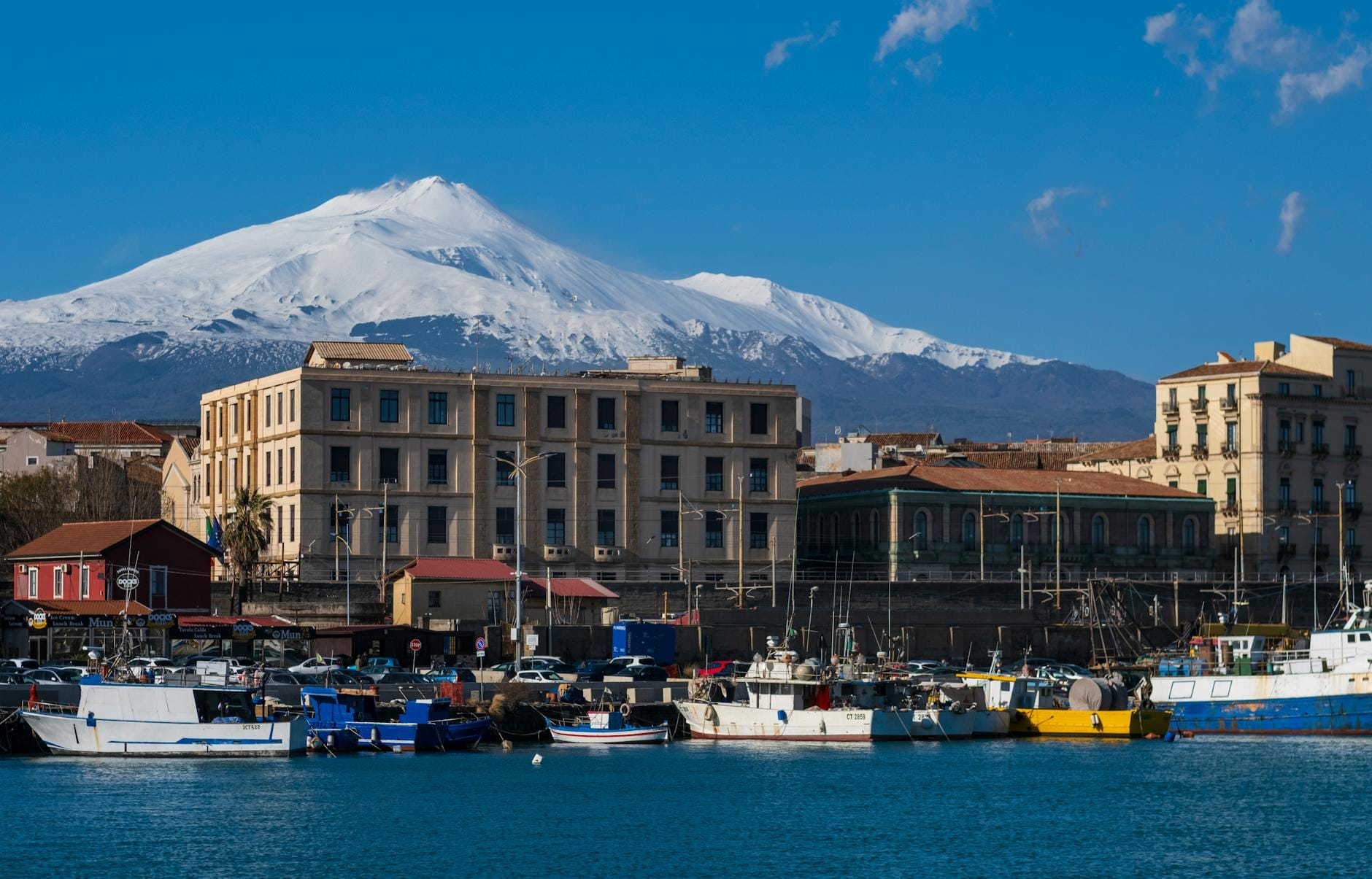 Küstenansicht von Catania mit Hafen, historischen Gebäuden und dem schneebedeckten Ätna im Hintergrund unter einem klaren blauen Himmel.