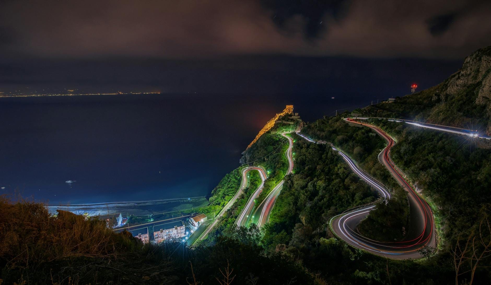 Nachtansicht einer kurvenreichen Küstenstraße an einem steilen Hang in Sizilien, mit Lichtspuren und dem Meer darunter.