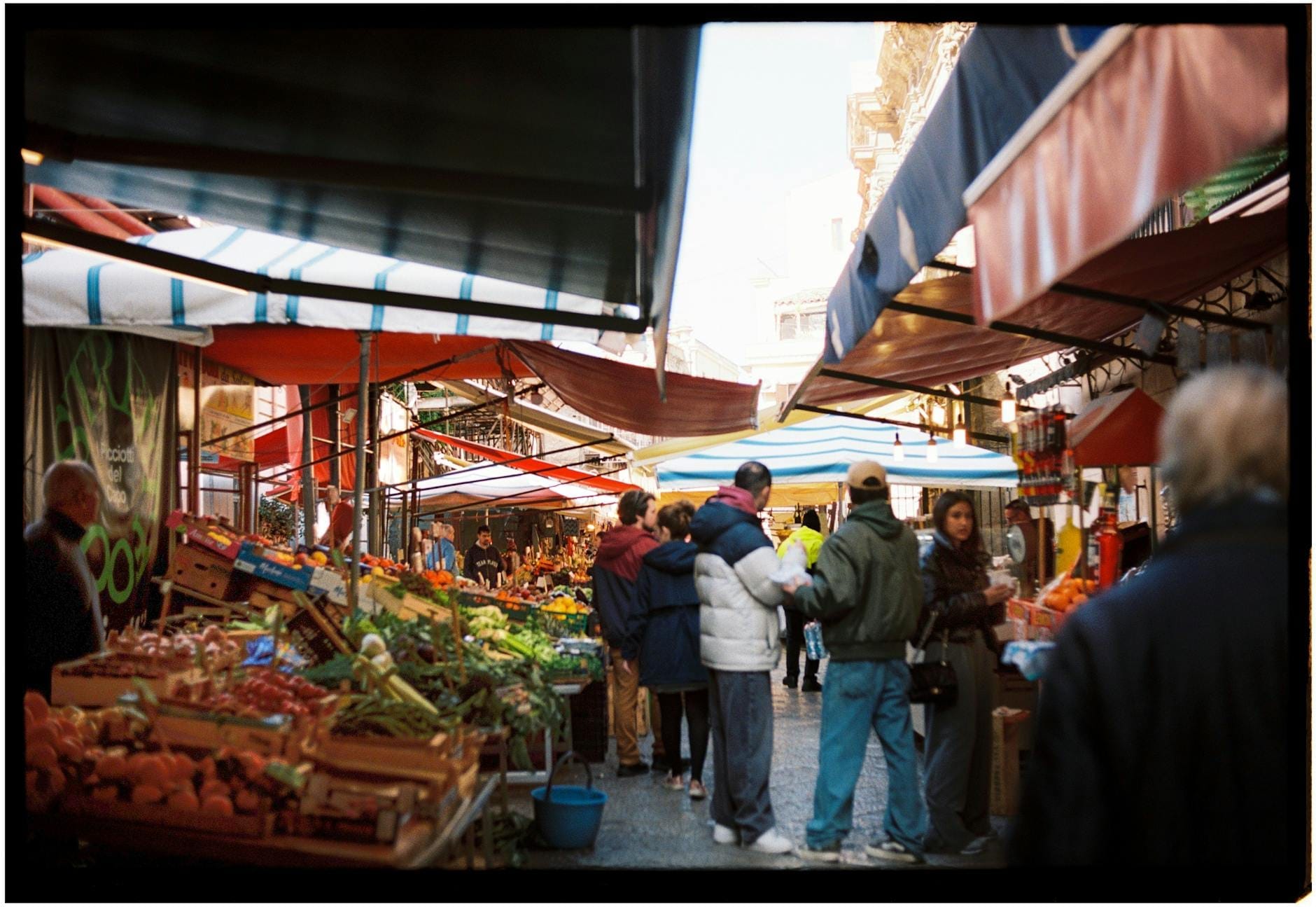 Belebter sizilianischer Freiluftmarkt mit bunten Markisen, Ständen mit frischen Produkten und Menschen beim Einkaufen und Plaudern in einer lebhaften Straßenszene.