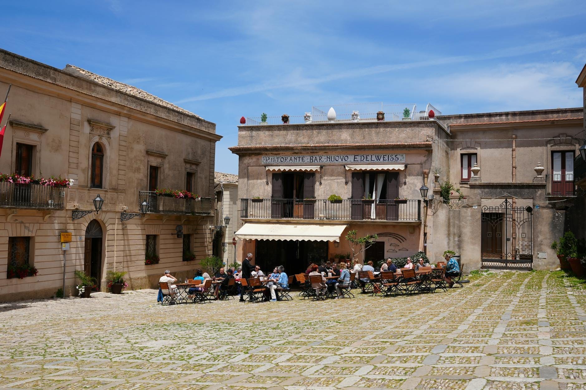 Volle Außentische am Bar Vitelli auf einer sonnenbeschienenen Piazza in Savoca, Sizilien, mit historischen Steingebäuden im Hintergrund.