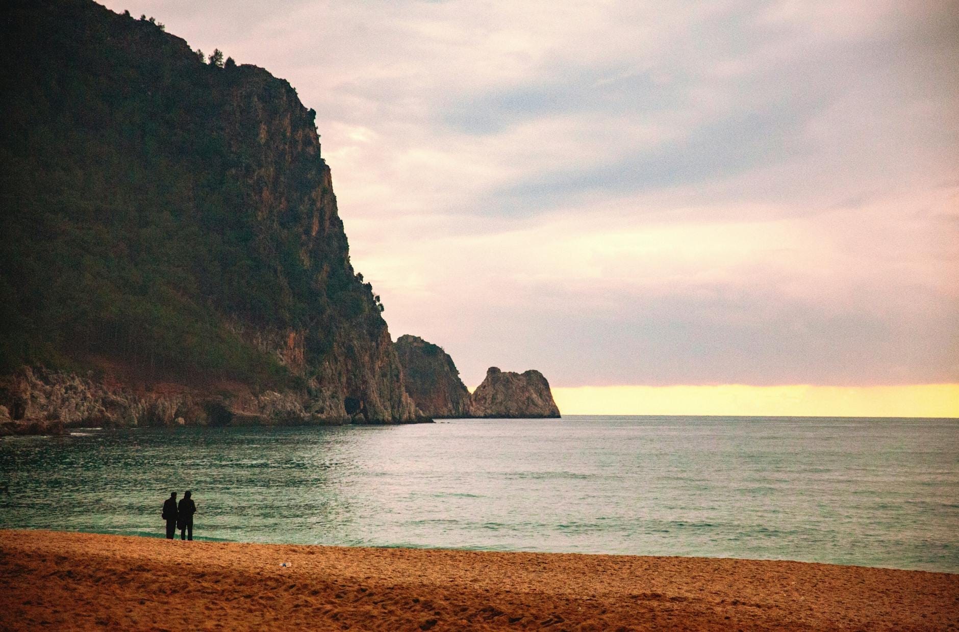 Ein Paar steht zusammen bei Sonnenuntergang an einem Sandstrand und blickt auf das Meer, während im Hintergrund dramatische Felsenklippen aufragen.