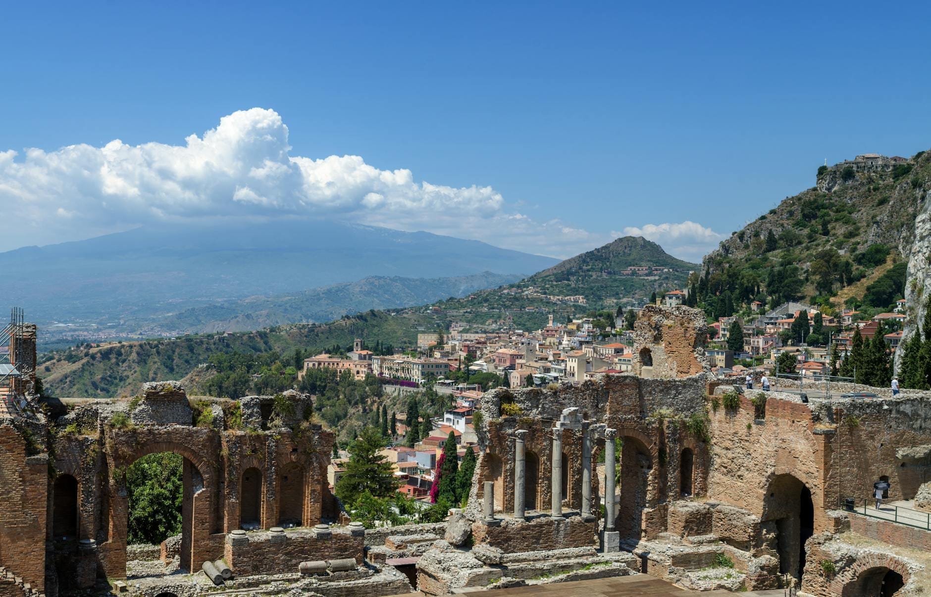 Panoramablick auf das antike Griechische Theater von Taormina mit dem Ätna, der Hanglage der Stadt und blauem Himmel im Hintergrund.