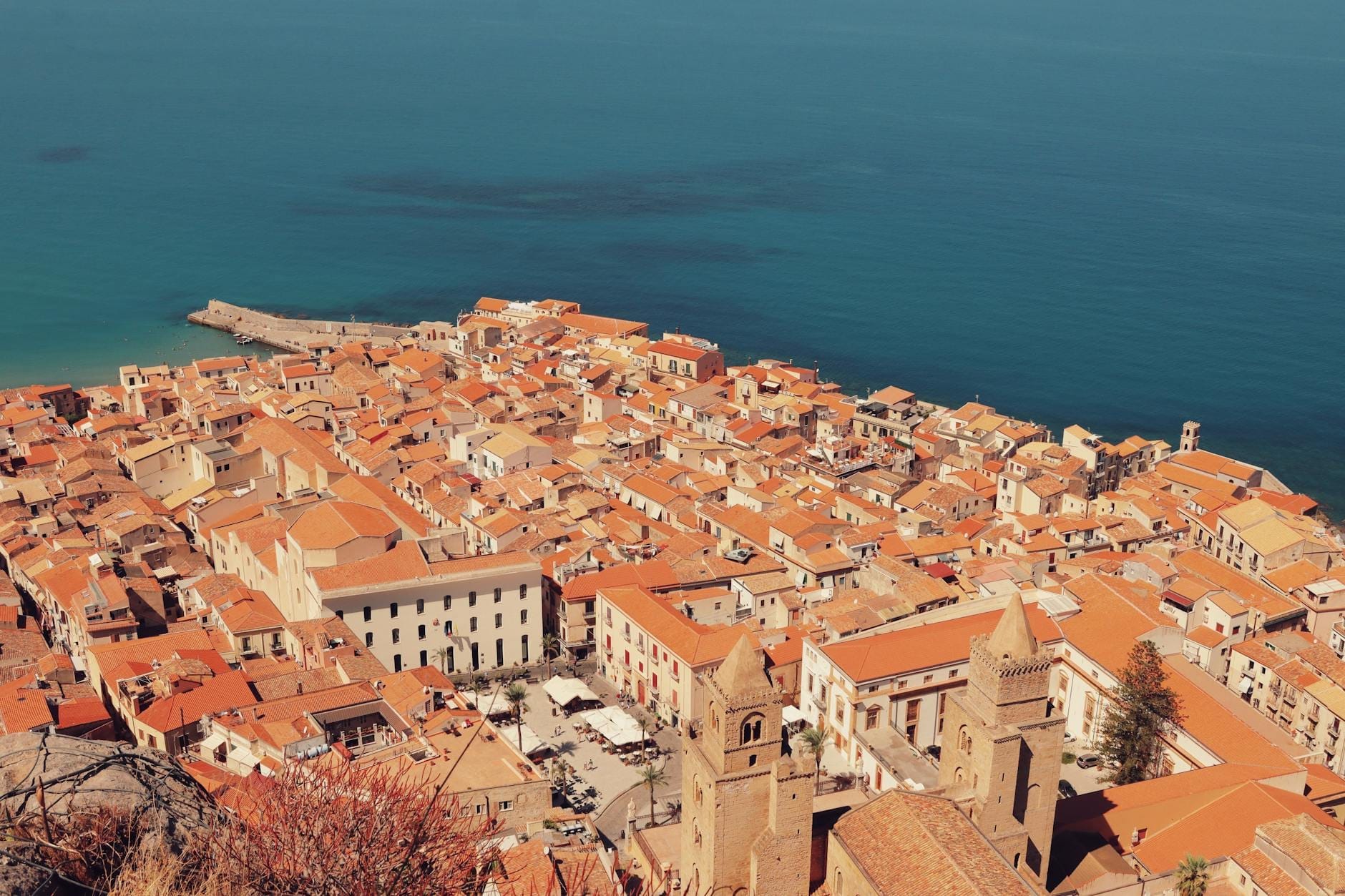 Klarer Blick über die sizilianische Küstenstadt Cefalù mit Terrakotta-Dächern, historischer Kathedrale und ruhigem blauem Meer unter mildem Sonnenhimmel.