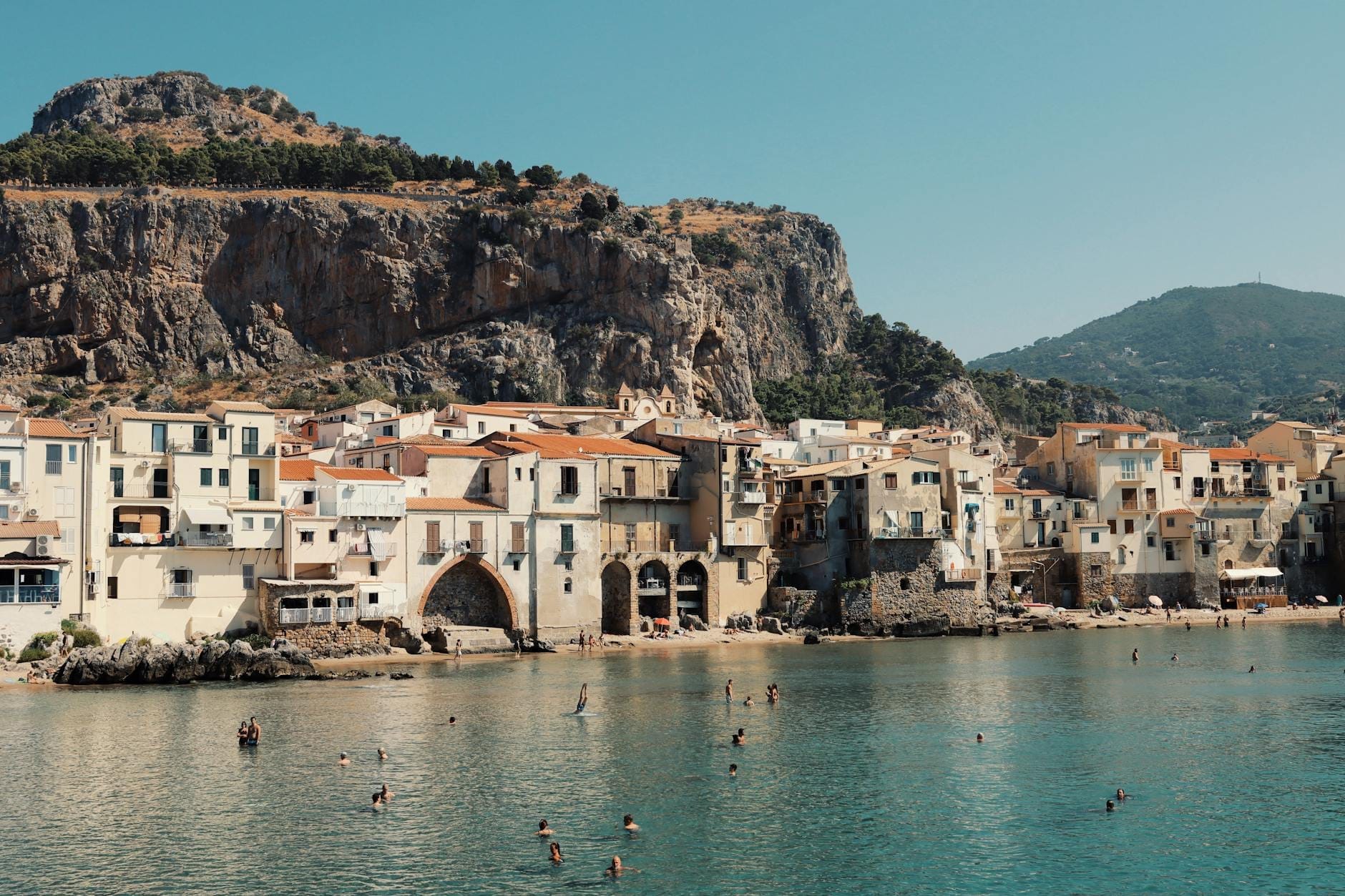 Sizilianische Küstenstadt mit historischen Gebäuden und Klippen, Menschen schwimmen im türkisfarbenen Meer unter einem klaren, sonnigen Sommerhimmel.