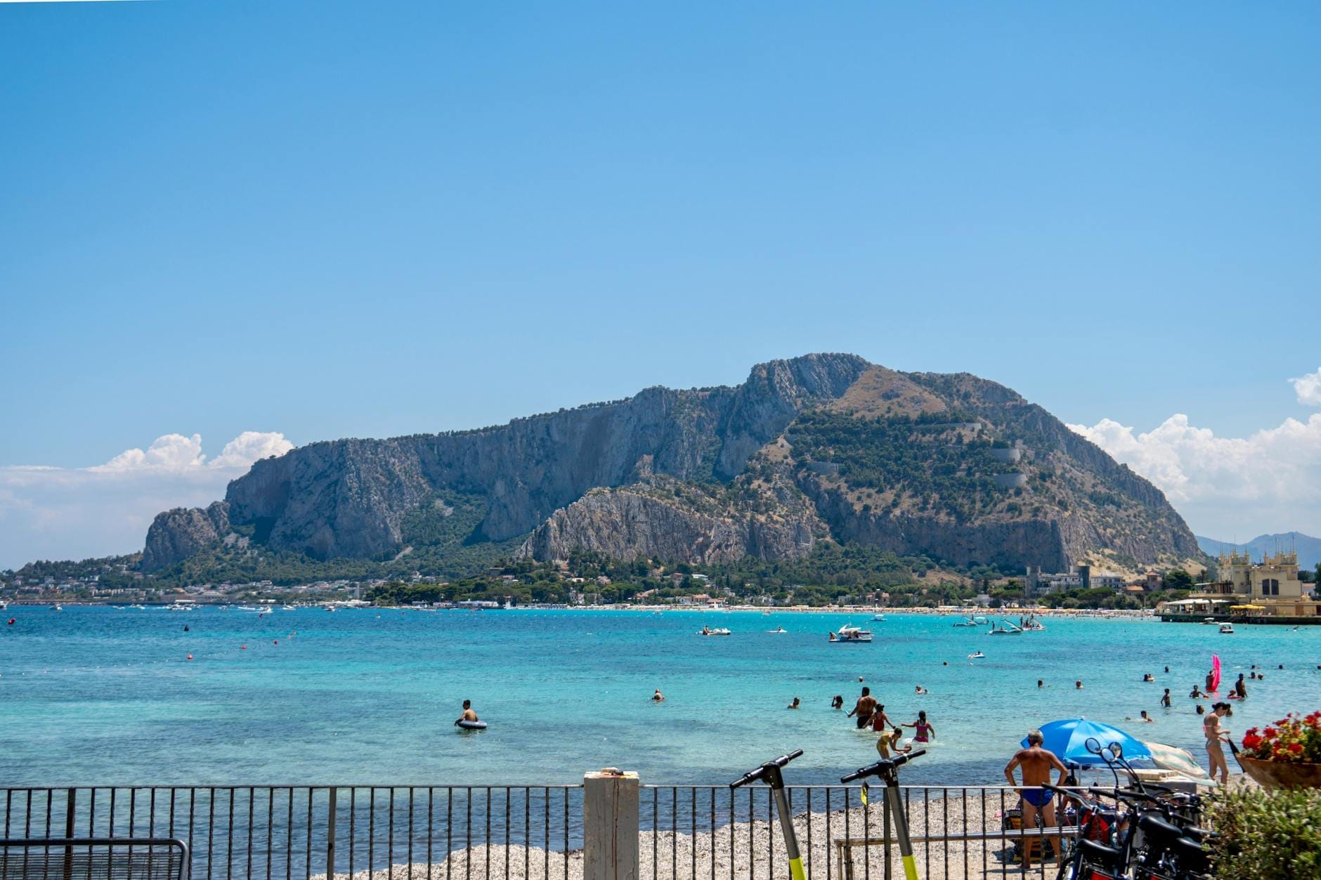 Überfüllter sizilianischer Strand mit türkisfarbenem Wasser, schwimmenden Menschen, Sonnenschirmen und einer dramatischen felsigen Bergkulisse unter einem sonnigen blauen Himmel.
