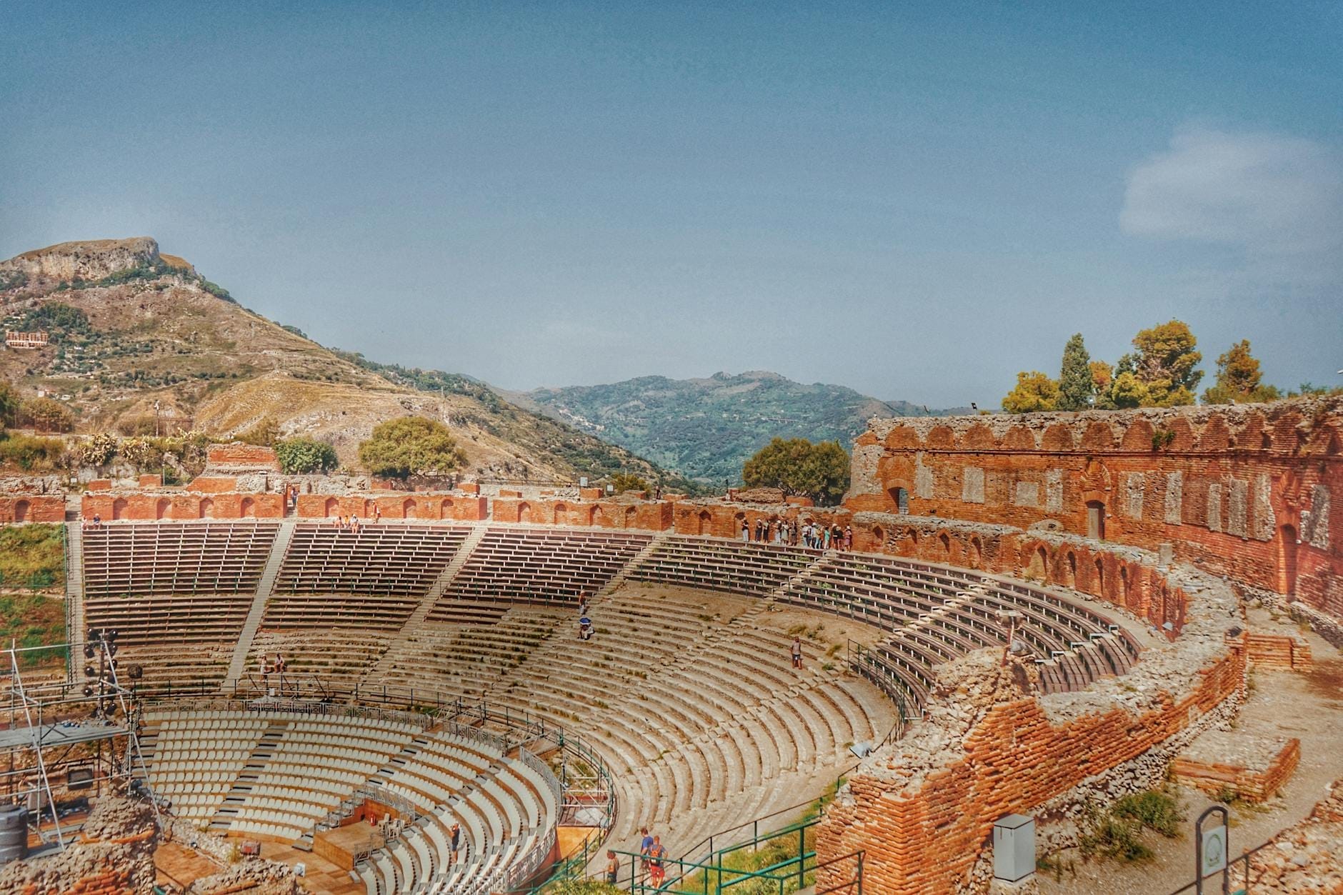 Weite Aussicht auf das antike Teatro Greco di Taormina mit den umliegenden Hügeln und blauem Himmel, ein ikonischer Veranstaltungsort für Siziliens Sommerfestivals und Events.