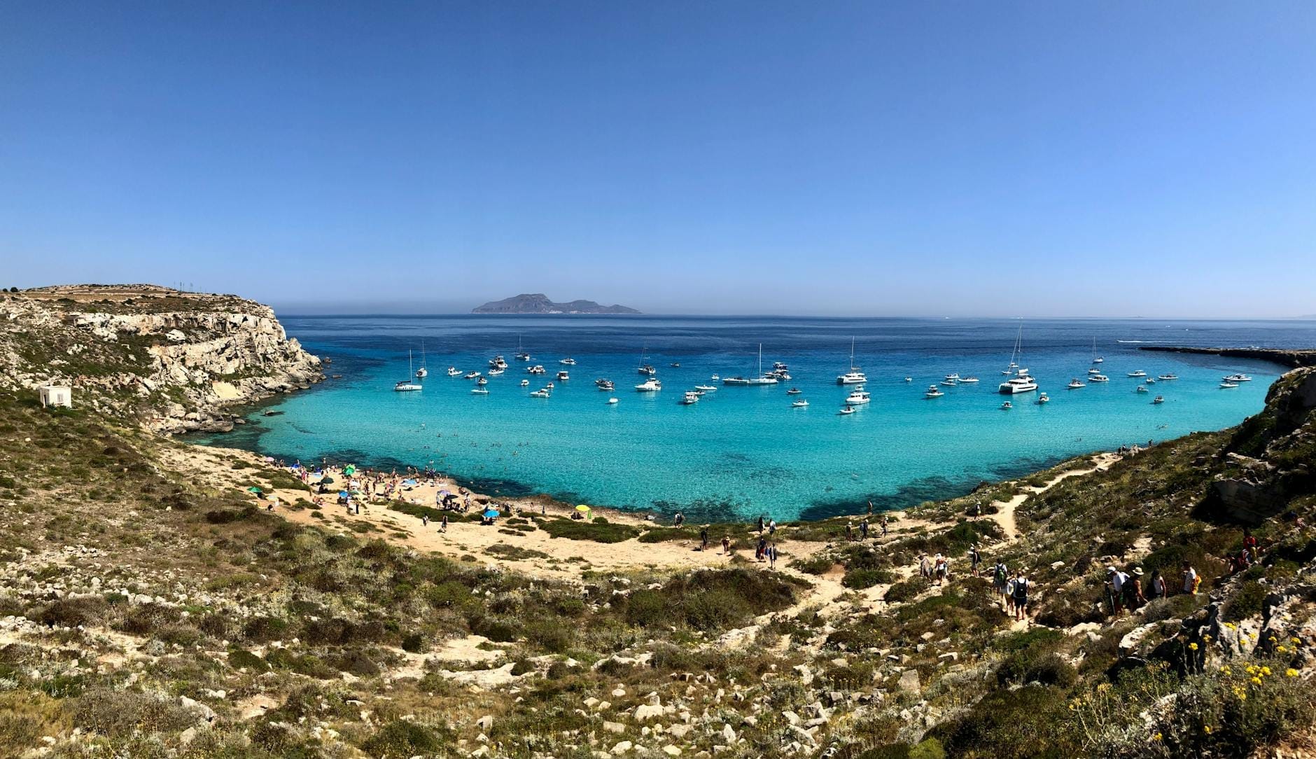 Panoramablick auf eine Bucht mit kristallklarem türkisfarbenem Wasser, vor Anker liegenden Booten, felsiger Küste und einem ruhigen Sandstrand an einem sonnigen Tag.