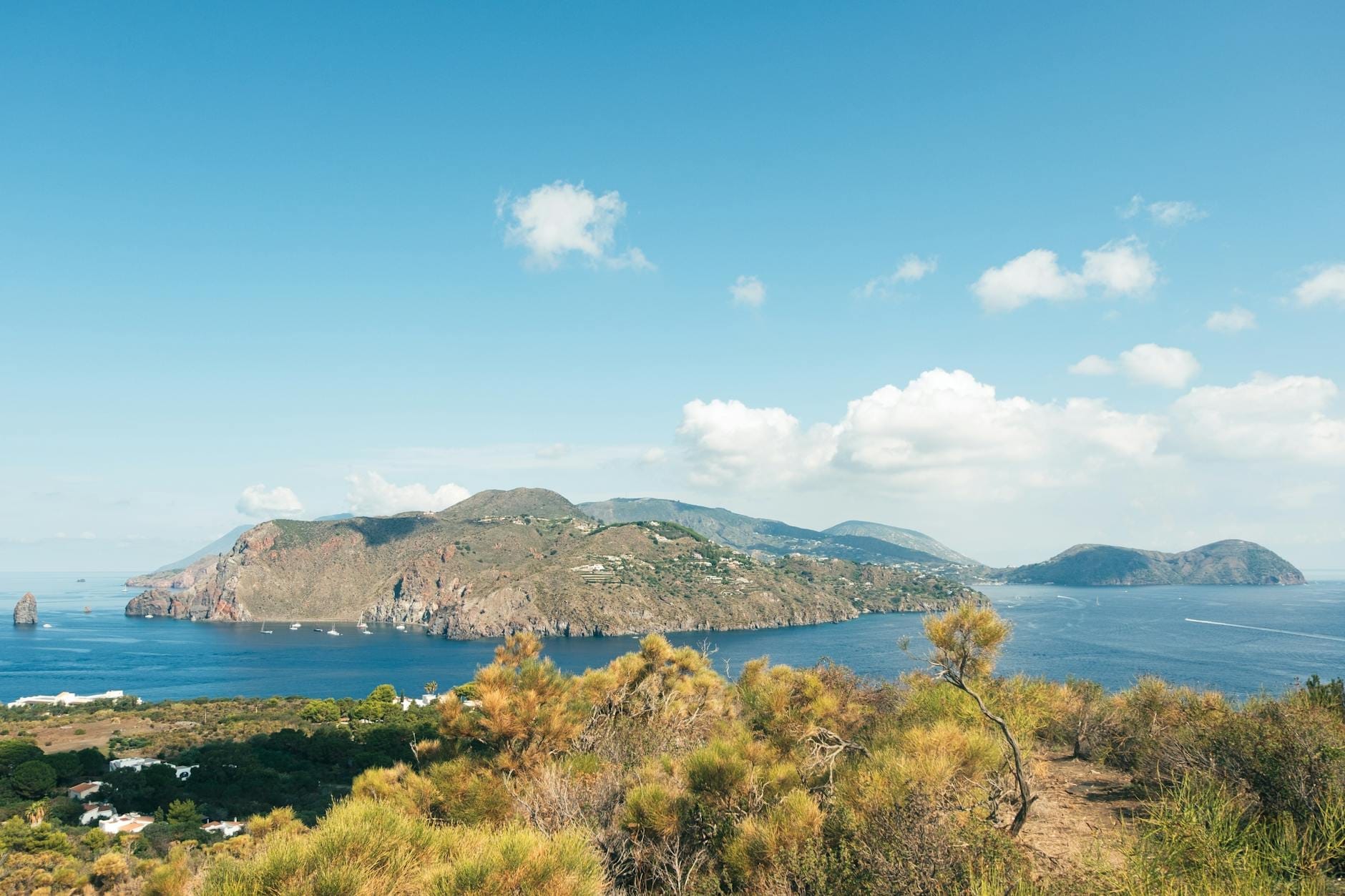 Panoramablick auf eine vulkanische Äolische Insel, umgeben vom tiefblauen Meer, fernen Inselchen, vereinzelten Wolken und mediterraner Vegetation im Vordergrund.