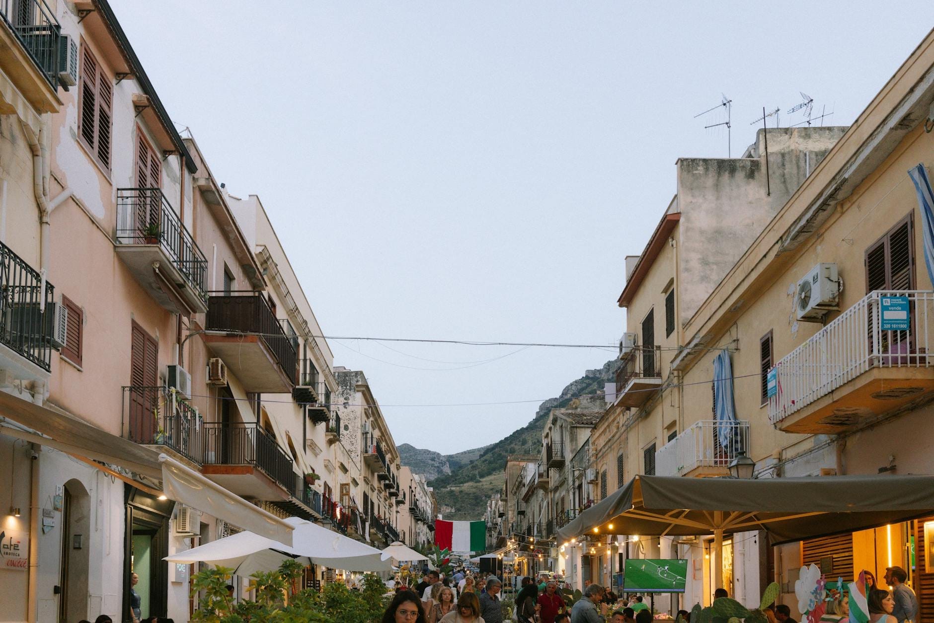 Straßenansicht in Sizilien mit spazierenden Menschen, bunten Gebäuden, Marktständen und Bergen im Hintergrund in der Abenddämmerung.
