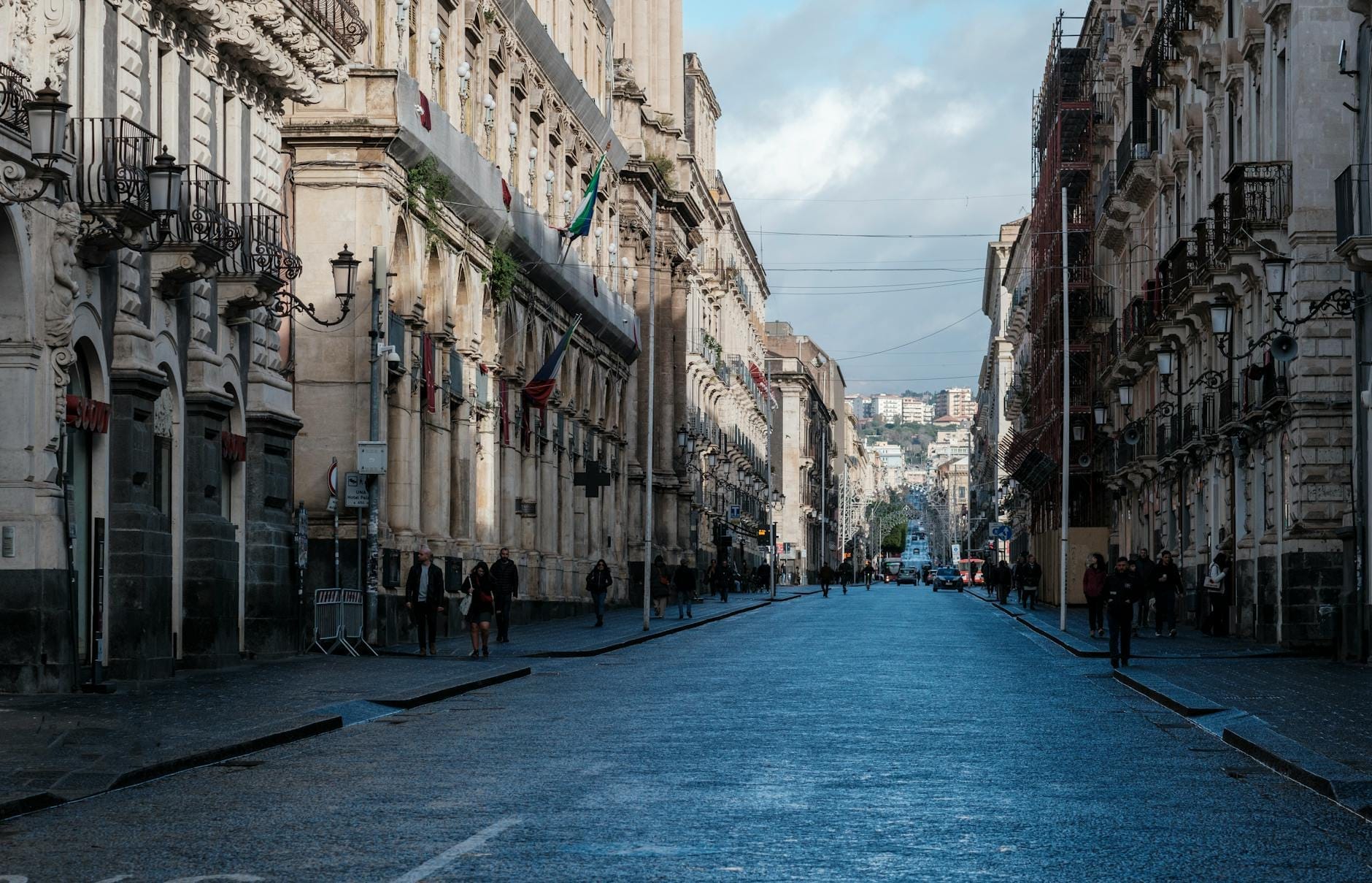Eine breite Straße in einer historischen sizilianischen Stadt mit prächtigen Barockgebäuden und Spaziergängern unter einem blauen Himmel mit vereinzelten Wolken.