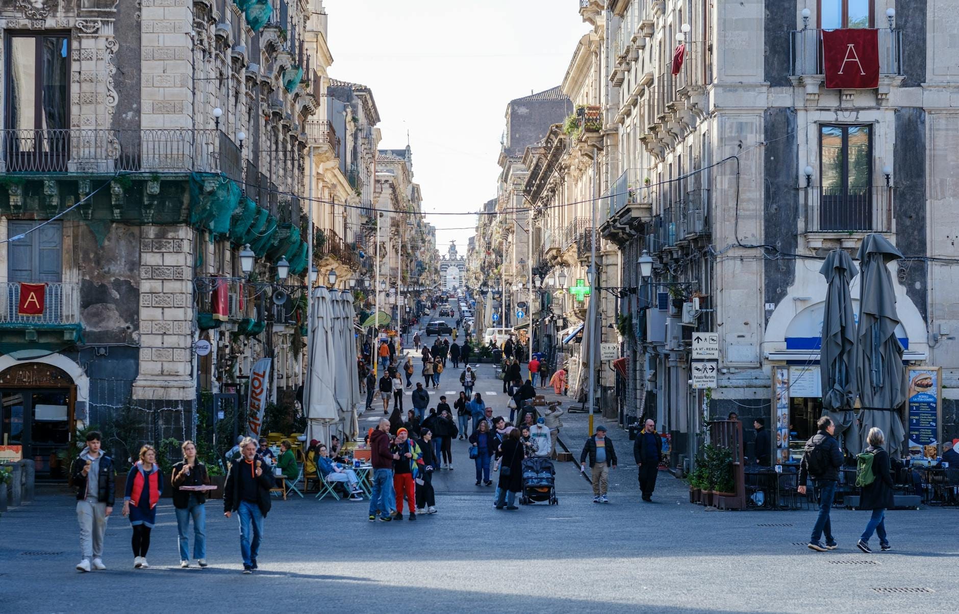 Belebte sizilianische Stadtstraße mit Fußgängern, historischen Steingebäuden auf beiden Seiten und einer einladenden, lebhaften Atmosphäre an einem sonnigen Tag.
