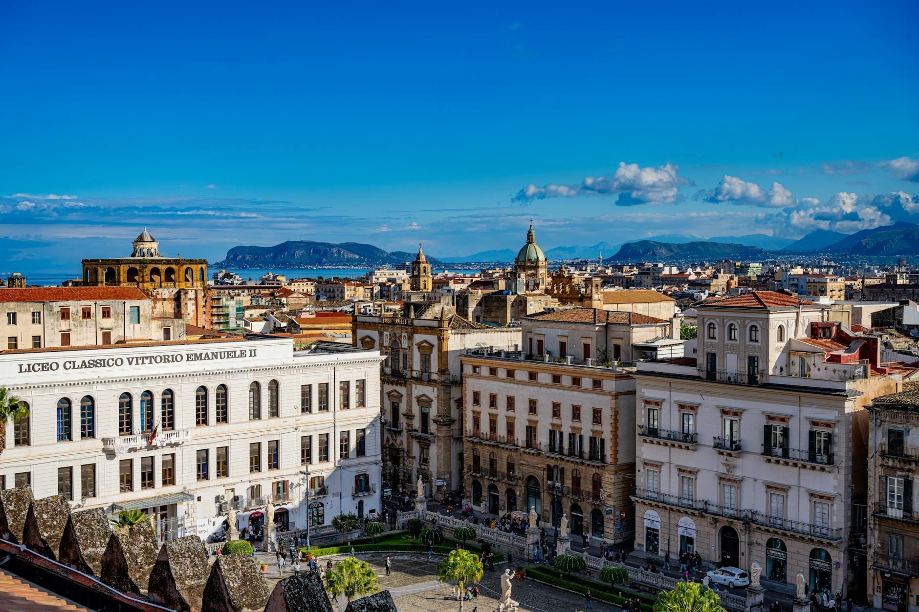 Panoramablick auf das historische Zentrum von Palermo mit klassischen Gebäuden, Kirchenkuppeln und fernen Bergen unter blauem Himmel.