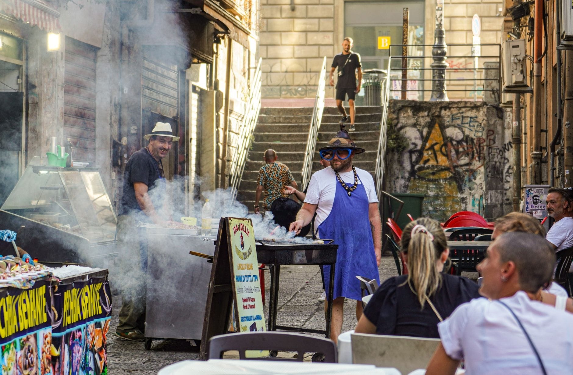 Street-Food-Händler grillen in einer lebhaften sizilianischen Gasse, während Einheimische und Touristen in der Nähe sitzen – rauchige Atmosphäre, bunte, lebendige Marktszene.
