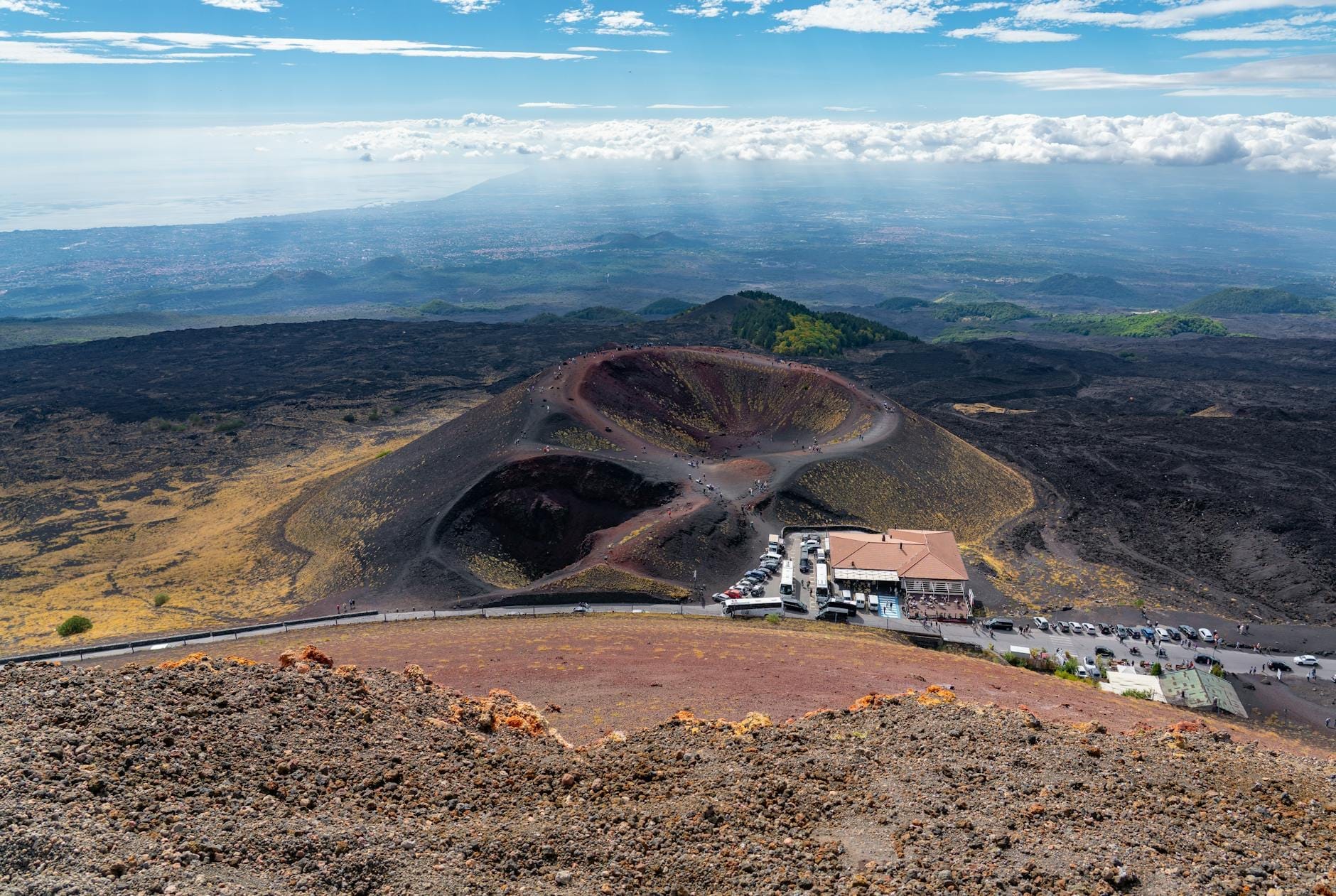 Weitblick auf die Vulkankrater des Ätna mit einer Berghütte und gewundenen Pfaden, Vulkanlandschaft unter einem hellen, teils bewölkten Himmel.
