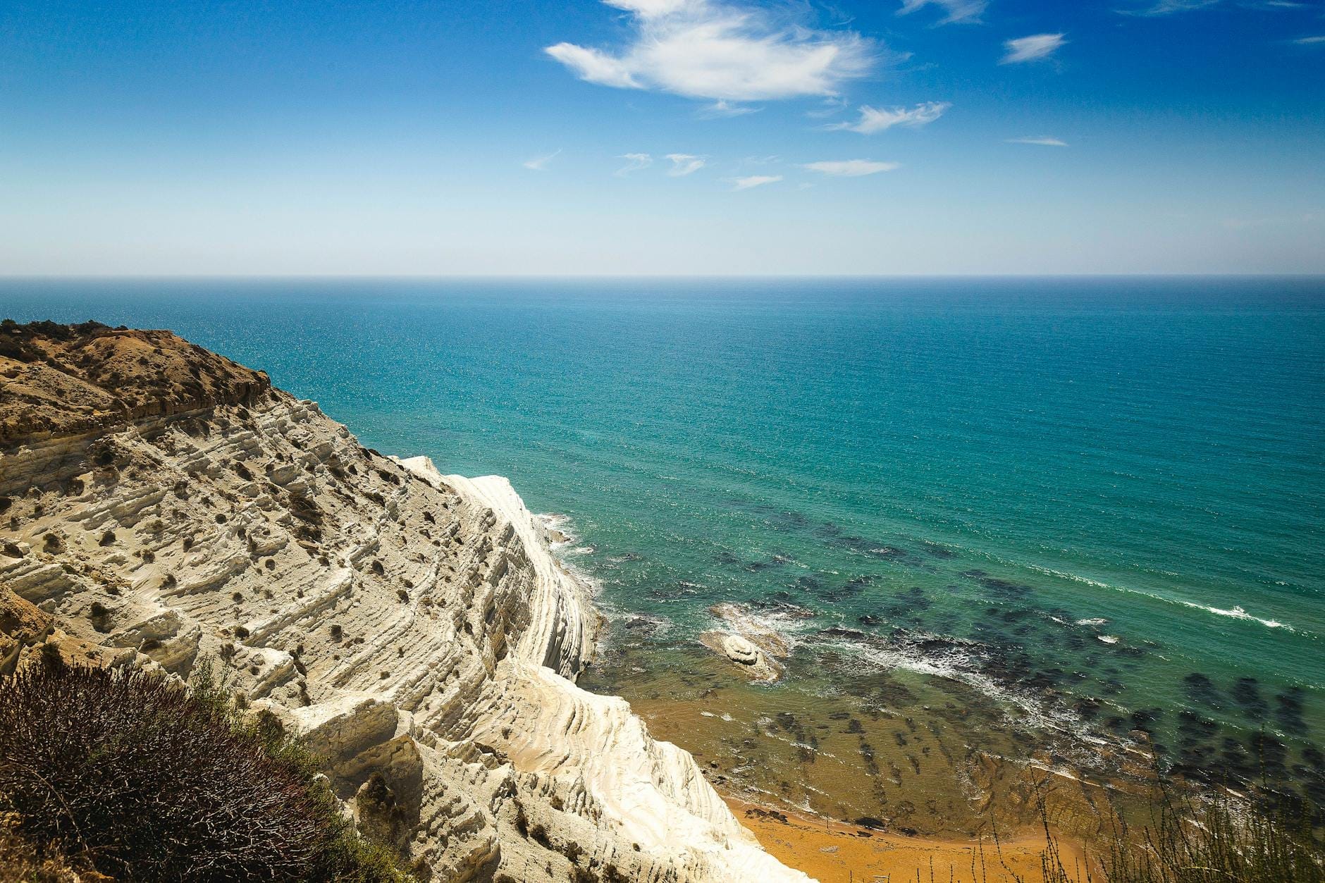 Weiße Kalksteinklippen treffen auf das türkisfarbene Meer unter einem blauen Himmel, mit etwas Gestrüpp im Vordergrund.
