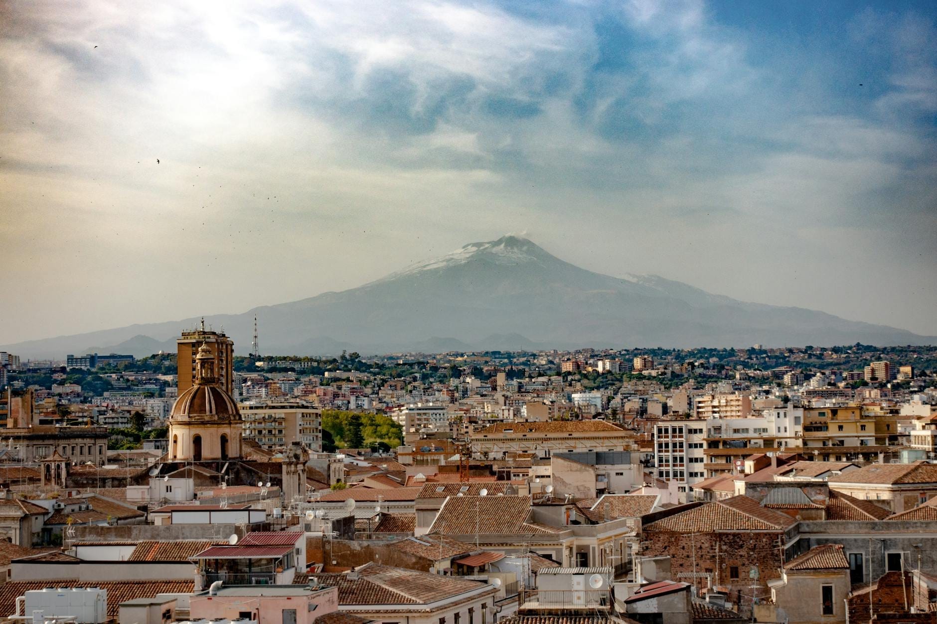 Wide view of a Sicilian city with traditional buildings in the foreground and the iconic Mount Etna volcano in the background under a dramatic sky.