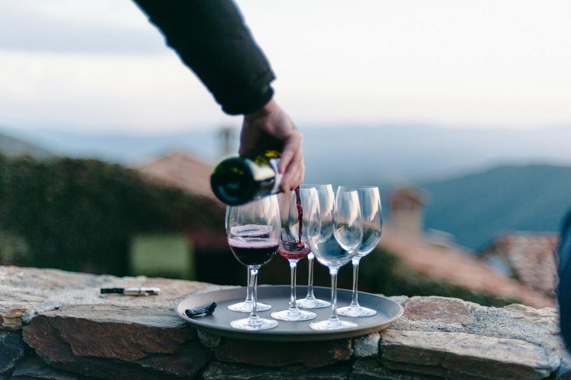 Wein wird in Gläser im Freien auf einer Steinmauer gegossen, mit einer hügeligen Landschaft im Hintergrund.