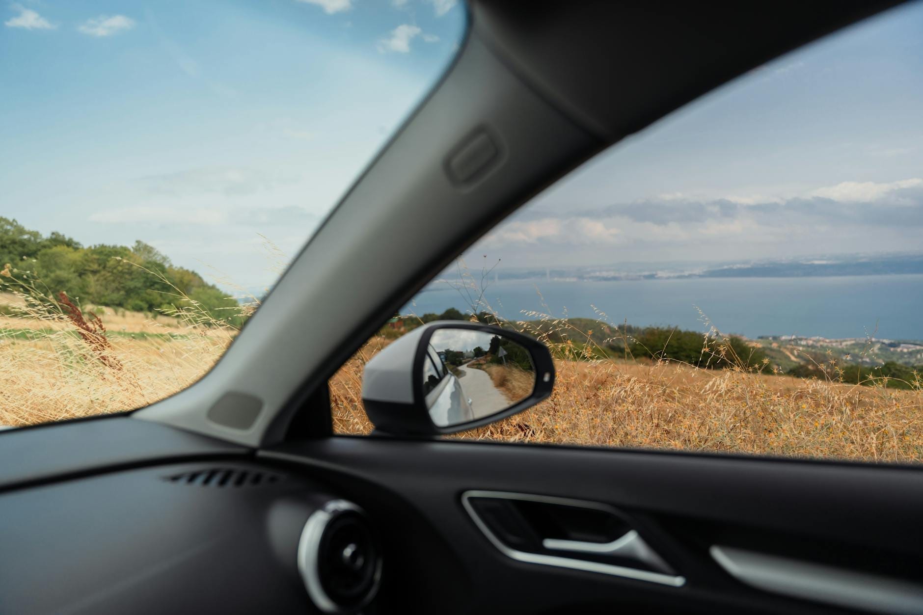 Blick aus dem Inneren eines Autos, das auf einer malerischen Küstenstraße auf dem Land fährt, mit Blick auf das Meer und die Hügel unter einem blauen Himmel.