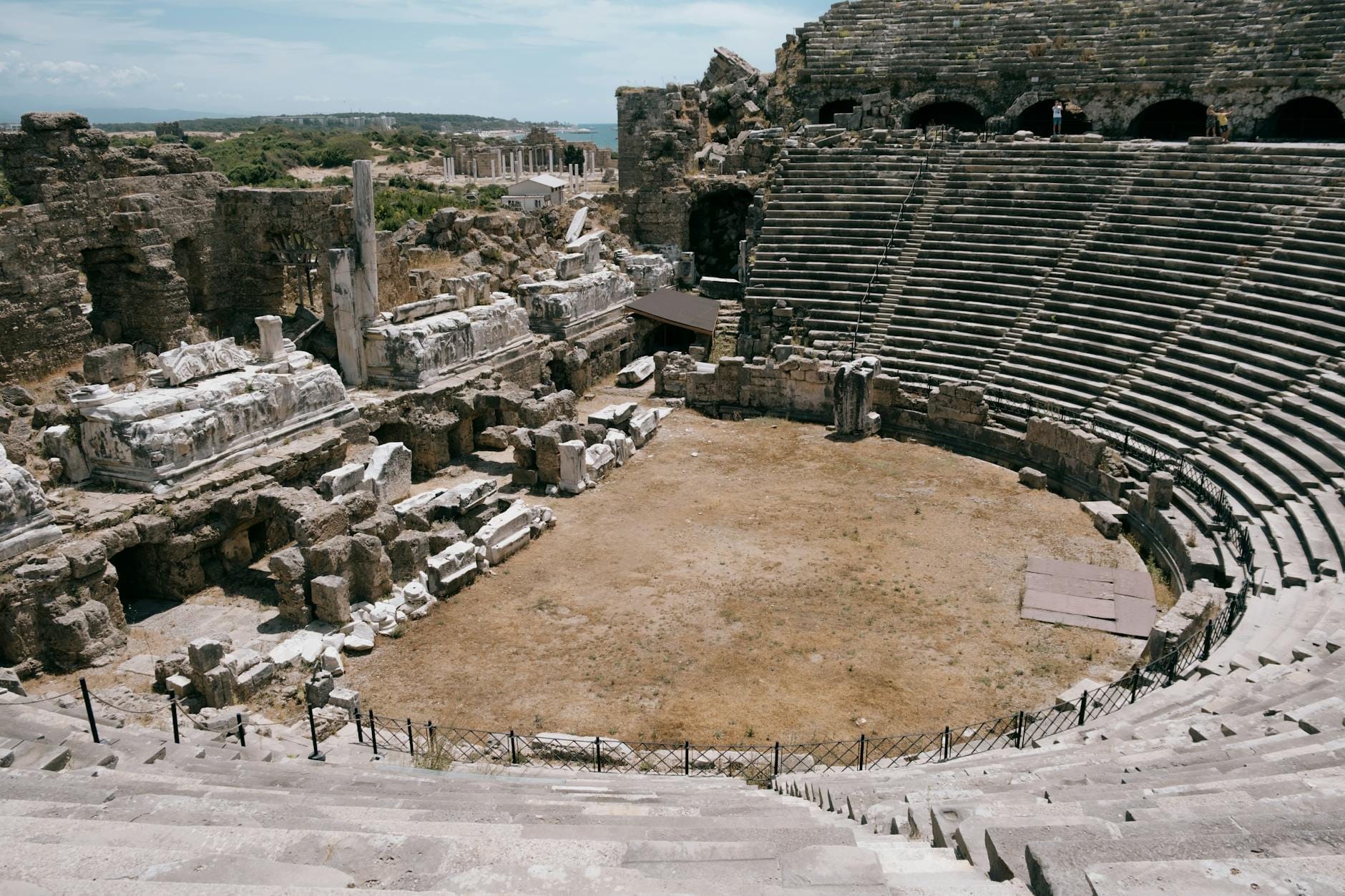 Vista grandangolare del Teatro Antico di Side con i gradoni in pietra e i resti parzialmente in piedi della scena romana, sotto un cielo luminoso — immagine perfetta per una copertina di viaggio.