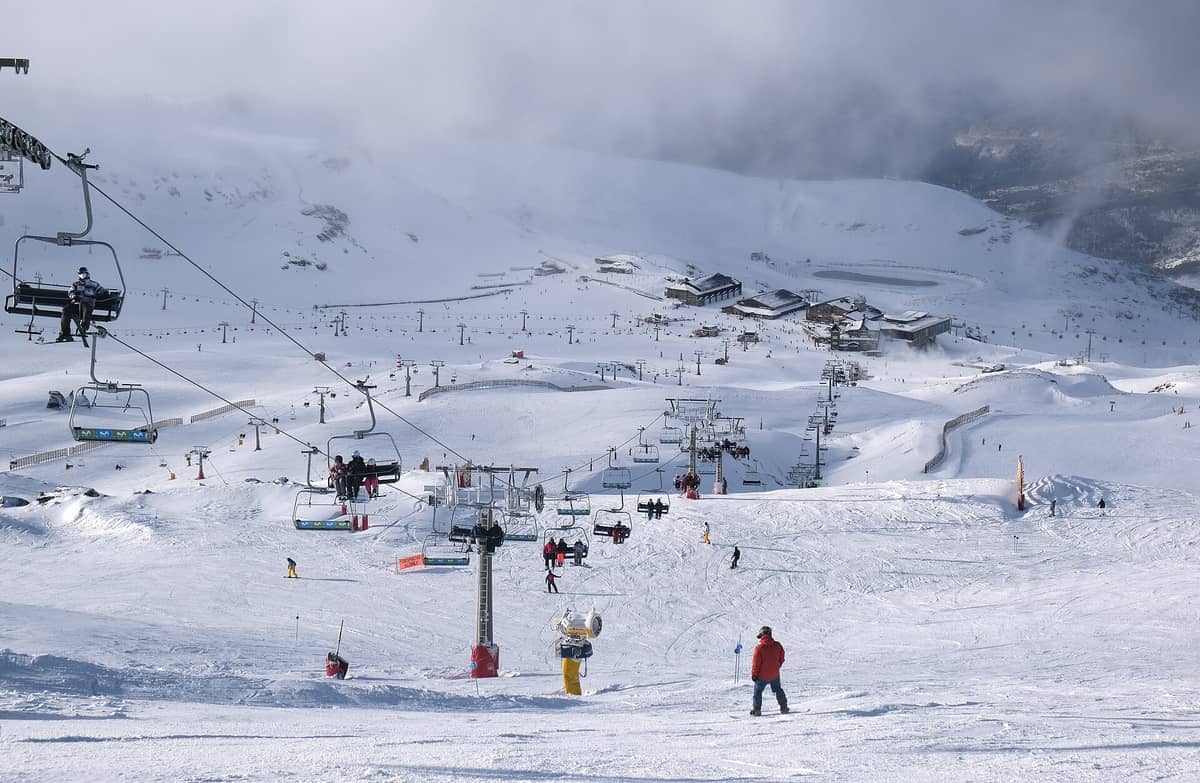 นักสกีโดยสารเก้าอี้ลอยฟ้าและลื่นไถลลงมาตามลาดเขาที่ปกคลุมด้วยหิมะที่ Sierra Nevada Ski and Mountain Resort โดยมีอาคารของรีสอร์ตและภูเขาที่ปกคลุมด้วยหมอกเป็นฉากหลัง