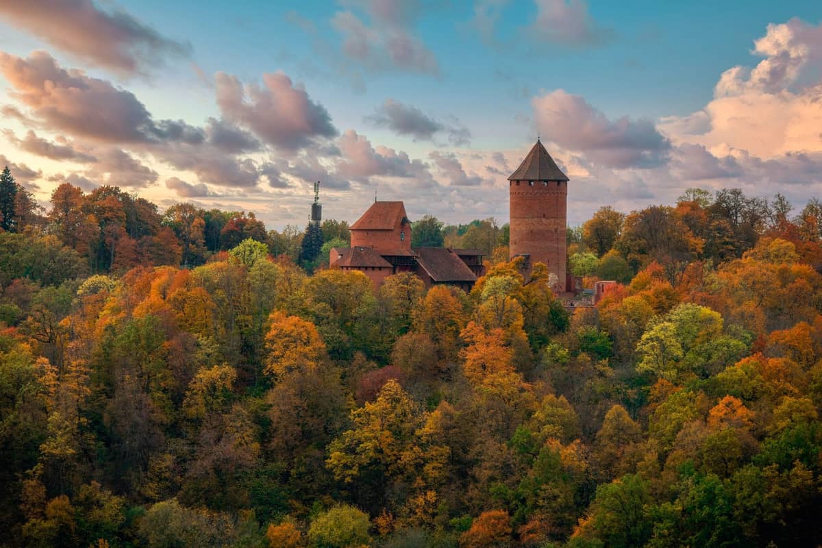 Luftaufnahme der Burg Turaida, umgeben von einem bunten Herbstwald in Sigulda, mit dramatischen Wolken und blauem Himmel darüber.