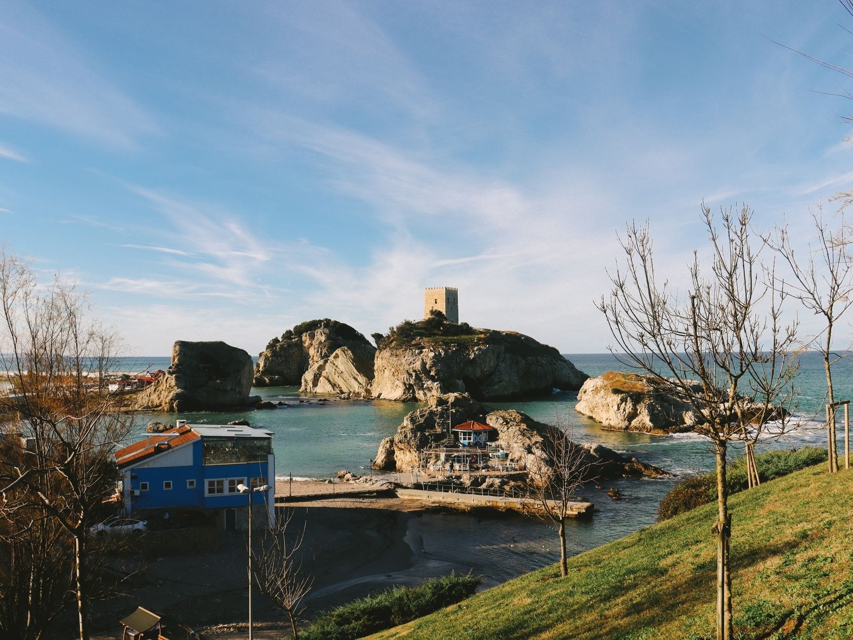 Isolotto roccioso con un castello medievale genovese circondato dalle acque turchesi del Mar Nero sotto un cielo luminoso, visto da una collina erbosa con edifici affacciati sul mare.