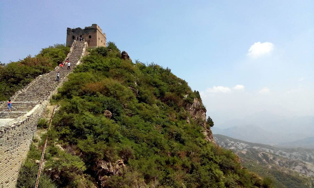 Steile, unrestaurierte Stufen führen zu einem verfallenen Wachturm auf der Simatai Chinesischen Mauer, umgeben von grünen Hügeln und einer beeindruckend rauen Landschaft.
