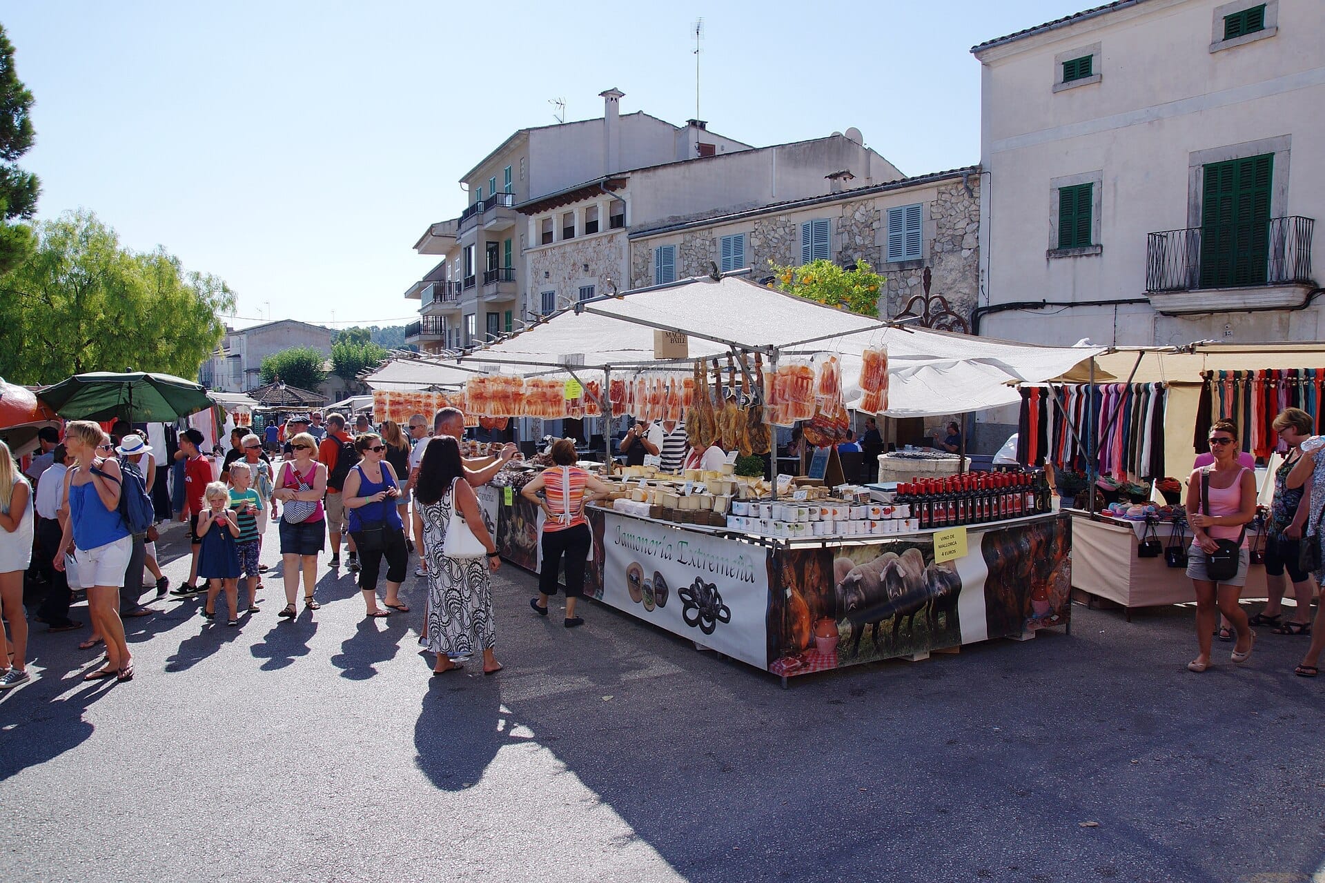 Le folle esplorano bancarelle all'aperto con prodotti locali, carni e artigianato al mercato del mercoledì di Sineu, con edifici in pietra tradizionali sullo sfondo.