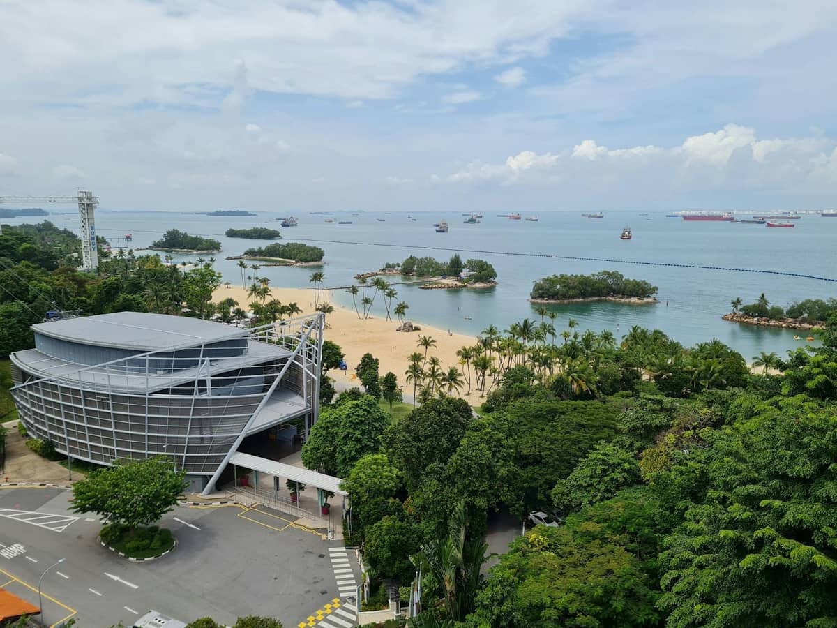Aerial view of Sentosa Island with sandy beaches, palm trees, a modern building, and ships anchored off the coast.