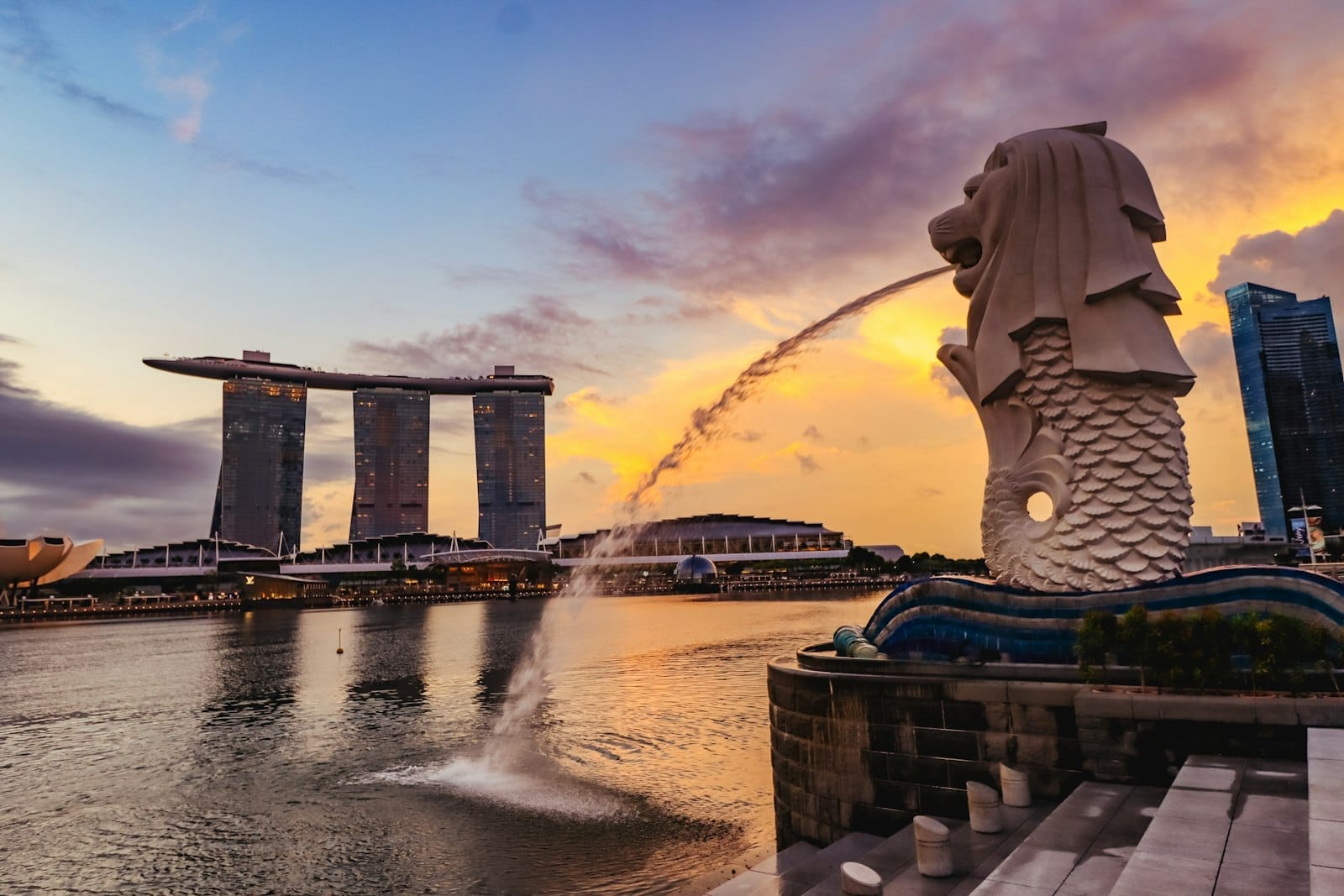 Estátua do Merlion em frente ao Marina Bay Sands ao pôr do sol, com céu dramático e skyline da cidade, convidando a explorar Singapura e escapadas próximas.