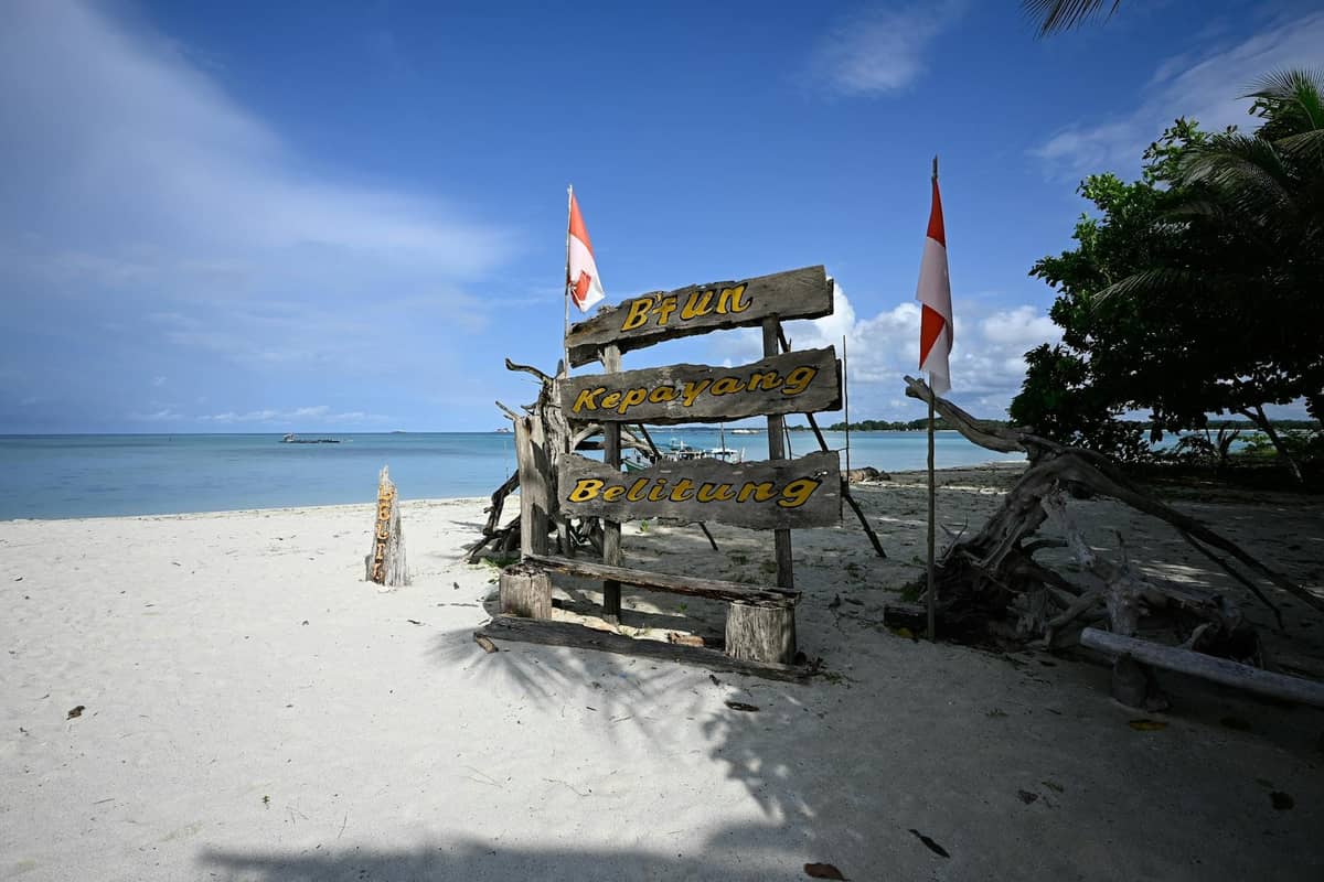 Sandy beach with wooden sign reading Bintan, Kepatang, Belitung and two Indonesian flags by the sea under blue sky