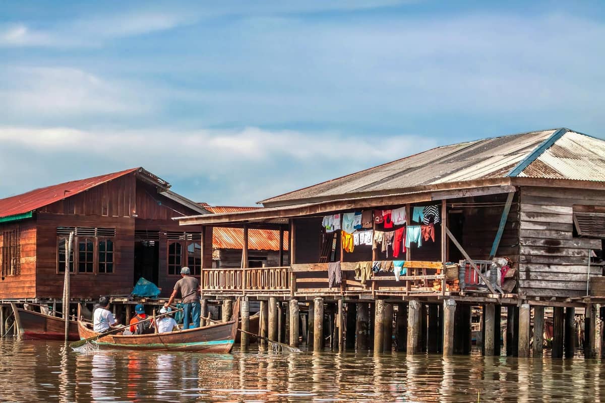 Wooden stilt houses over water with laundry hanging, a small boat with people in front, blue sky background.
