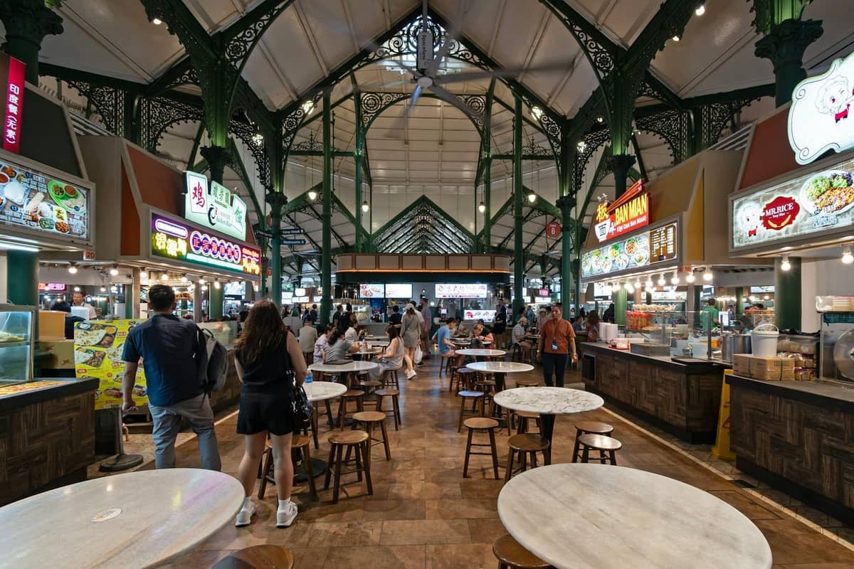 Interior of a bustling Singapore hawker centre with food stalls, shared tables, and people ordering and eating.