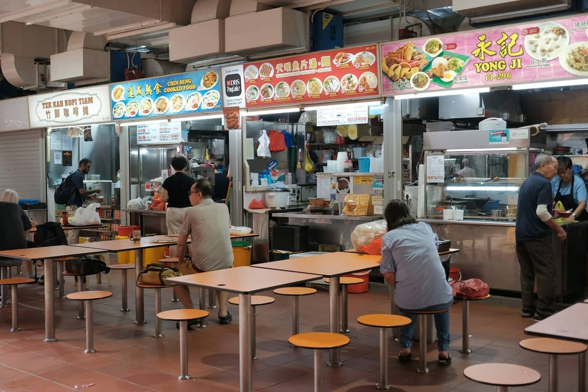 Interior of a Singapore hawker centre with food stalls, menus above, and people dining at communal tables.